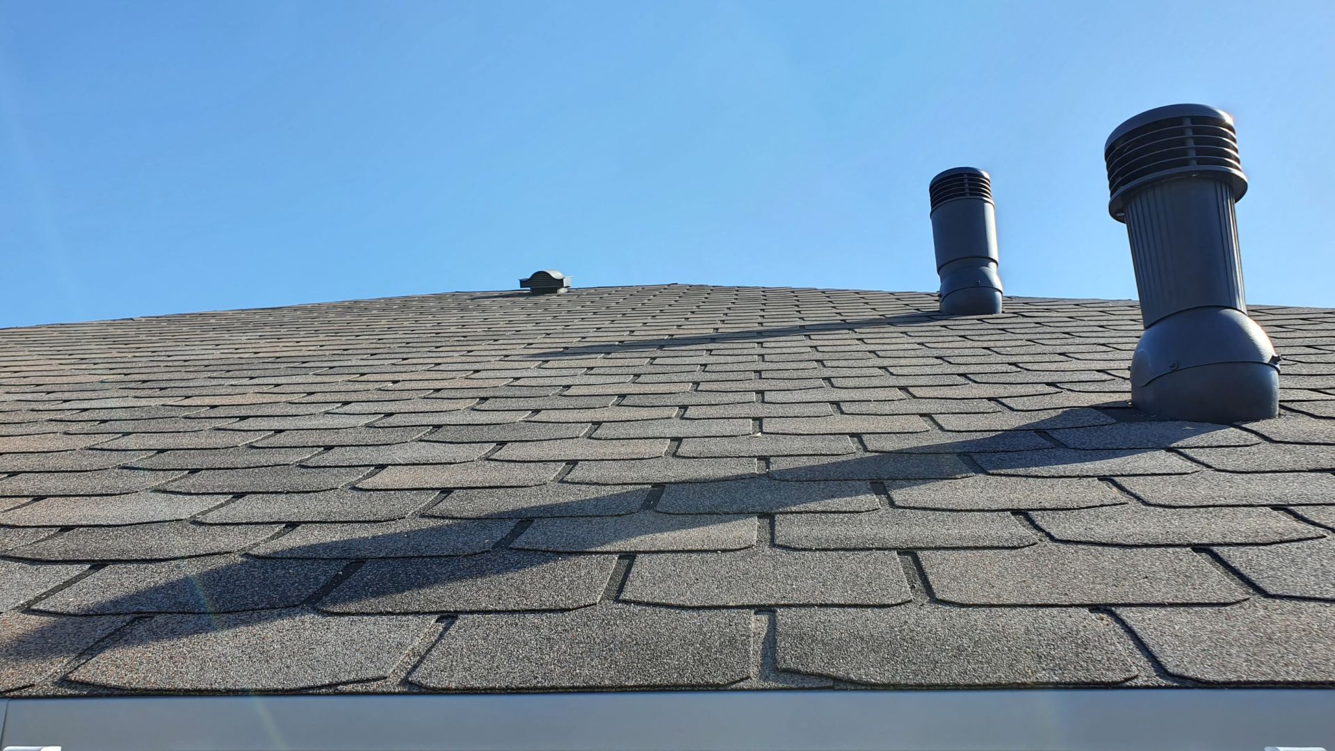 Gray asphalt shingle roof with two ventilation pipes against blue sky